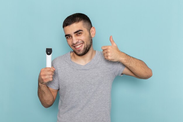 Vista frontal del hombre joven en camiseta gris con maquinilla de afeitar eléctrica sonriendo en la espuma de pelo masculino de barba azul afeitado