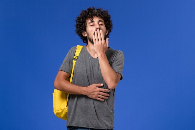 Vista frontal del hombre joven en camiseta gris con mochila amarilla bostezando en la pared azul