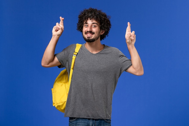 Vista frontal del hombre joven en camiseta gris con mochila amarilla cruzando los dedos sobre la pared azul