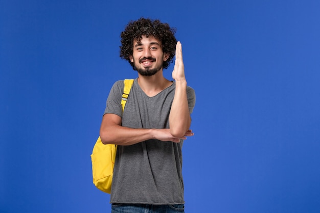 Vista frontal del hombre joven en camiseta gris con mochila amarilla levantando su mano sobre la pared azul
