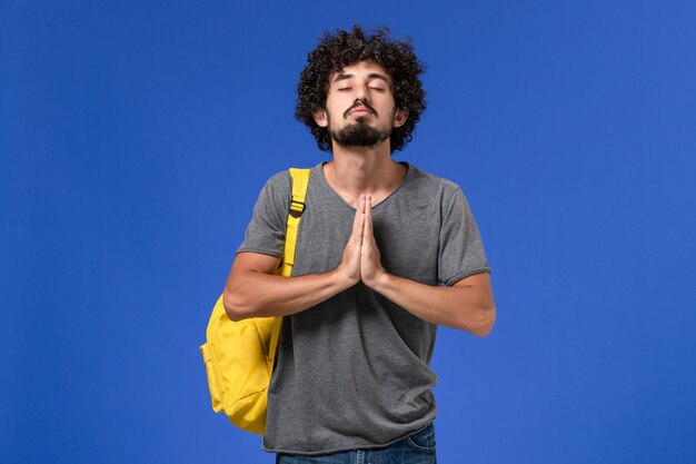 Vista frontal del hombre joven en camiseta gris con mochila amarilla orando en la pared azul