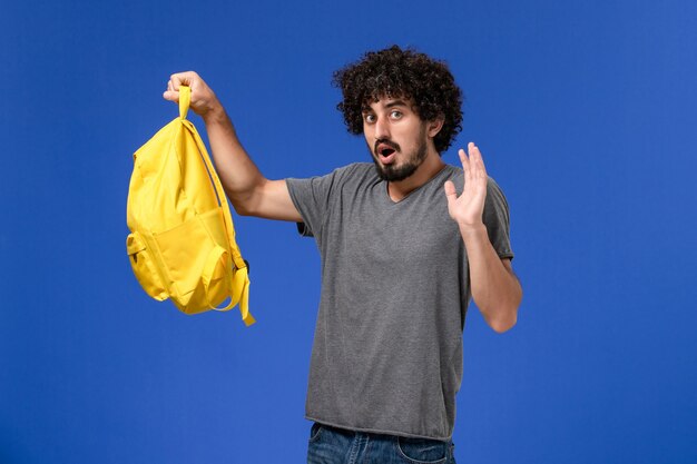 Vista frontal del hombre joven en camiseta gris con mochila amarilla en la pared azul