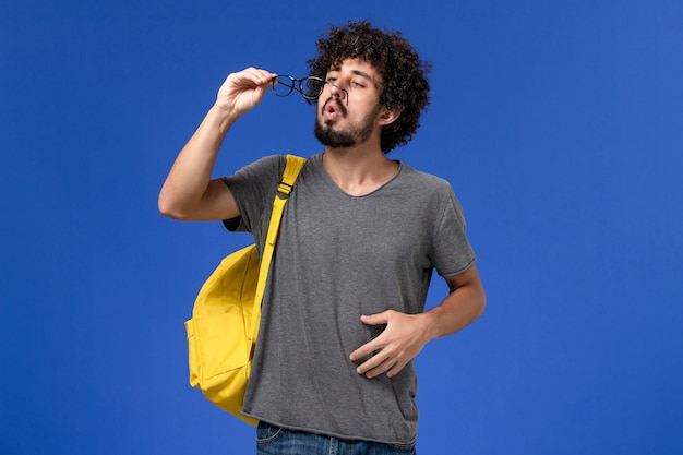 Vista frontal del hombre joven en camiseta gris con mochila amarilla en la pared azul