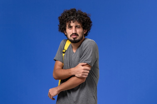 Vista frontal del hombre joven en camiseta gris con mochila amarilla en la pared azul