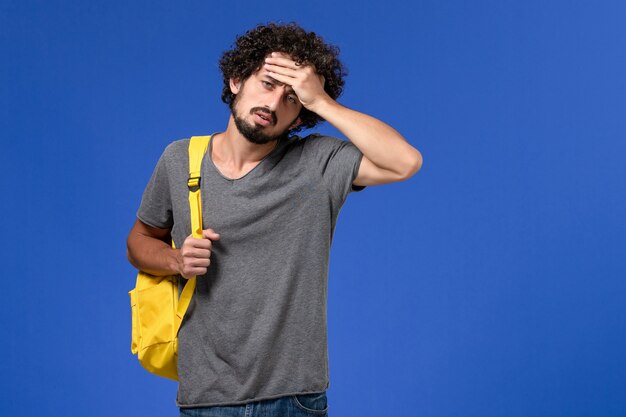 Vista frontal del hombre joven en camiseta gris con mochila amarilla en la pared azul