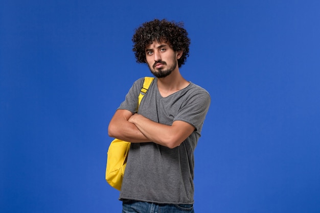 Vista frontal del hombre joven en camiseta gris con mochila amarilla pensando profundamente en la pared azul
