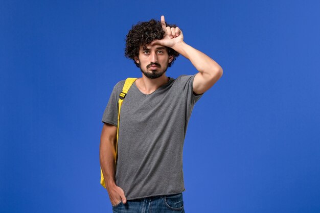 Vista frontal del hombre joven en camiseta gris con mochila amarilla posando en la pared azul claro