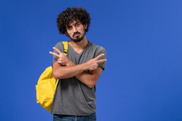 Vista frontal del hombre joven en camiseta gris con mochila amarilla posando en la pared azul