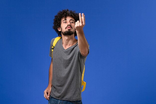 Vista frontal del hombre joven en camiseta gris con mochila amarilla posando en la pared azul