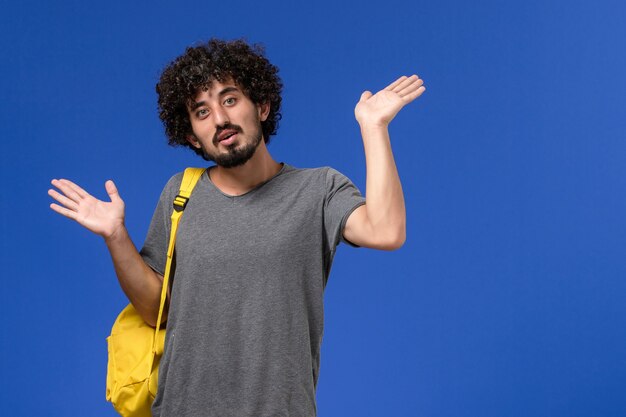 Vista frontal del hombre joven en camiseta gris con mochila amarilla posando en la pared azul