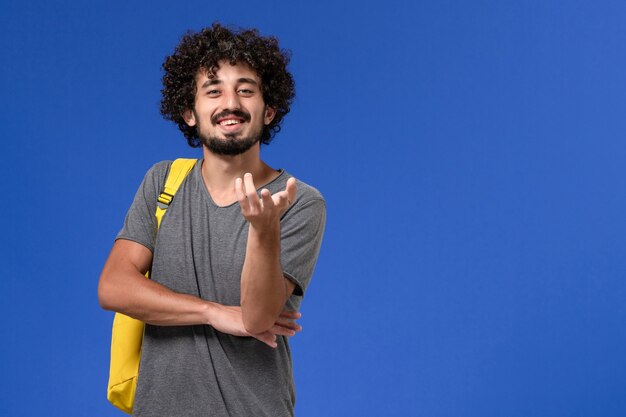 Vista frontal del hombre joven en camiseta gris con mochila amarilla posando con una sonrisa en la pared azul