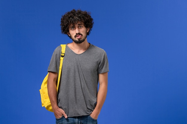 Vista frontal del hombre joven en camiseta gris con mochila amarilla solo de pie en la pared azul