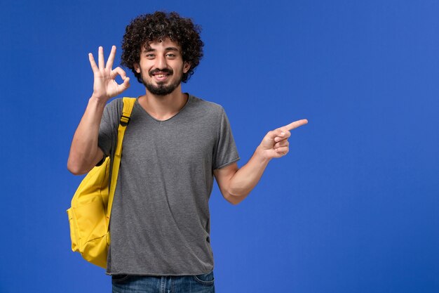 Vista frontal del hombre joven en camiseta gris con mochila amarilla sonriendo en la pared azul