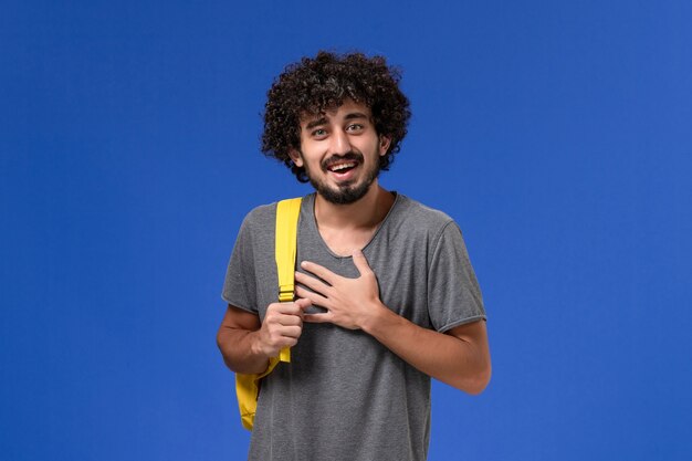 Foto gratuita vista frontal del hombre joven en camiseta gris con mochila amarilla sonriendo en la pared azul