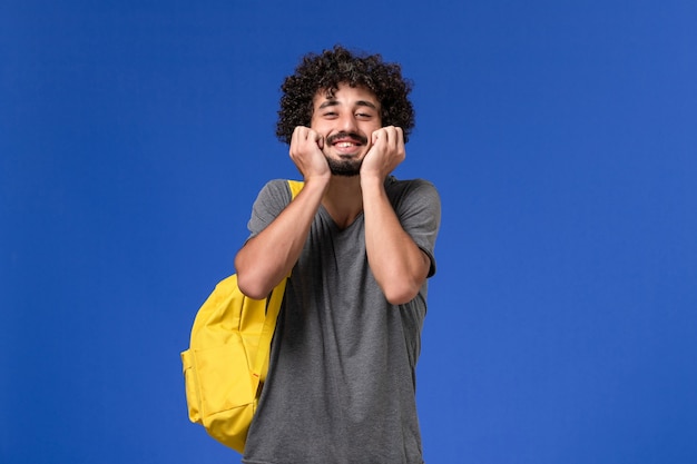 Vista frontal del hombre joven en camiseta gris con mochila amarilla sonriendo en la pared azul