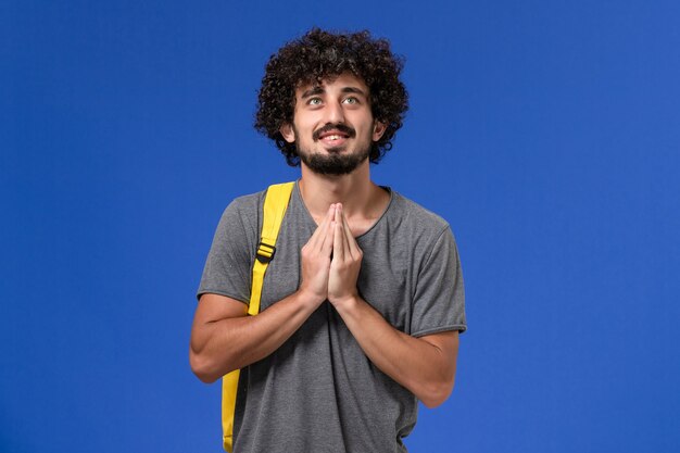 Vista frontal del hombre joven en camiseta gris con mochila amarilla sonriendo y rezando en la pared azul