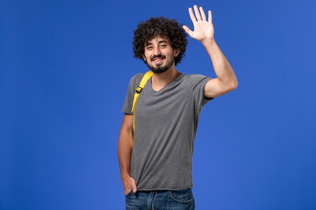 Vista frontal del hombre joven en camiseta gris con mochila amarilla sonriendo y saludando en la pared azul