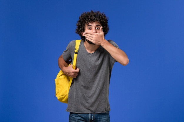 Vista frontal del hombre joven en camiseta gris con mochila amarilla tapándose la boca en la pared azul