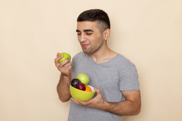 Vista frontal del hombre joven en camiseta gris con placa con frutas comiendo manzana en beige