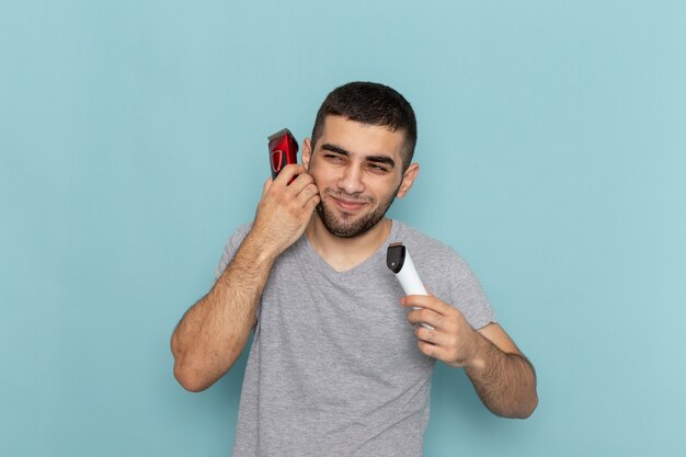 Vista frontal del hombre joven en camiseta gris sosteniendo dos maquinillas de afeitar eléctricas diferentes en azul helado