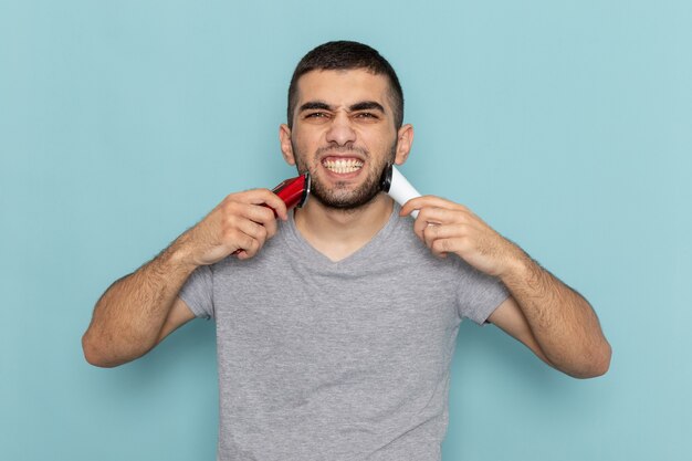 Foto gratuita vista frontal del hombre joven en camiseta gris sosteniendo dos maquinillas de afeitar eléctricas diferentes en el azul hielo