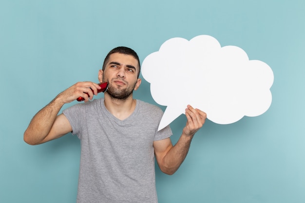 Vista frontal hombre joven en camiseta gris sosteniendo un gran cartel blanco y maquinilla de afeitar eléctrica en la pared azul helado barba masculina afeitadora de pelo