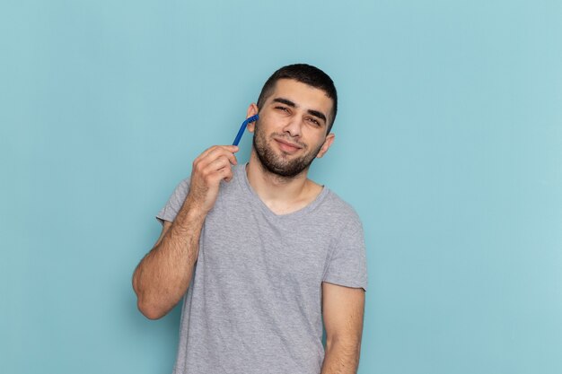 Vista frontal hombre joven en camiseta gris sosteniendo la navaja y afeitándose la cara en la barba de afeitar azul color de espuma de cabello masculino