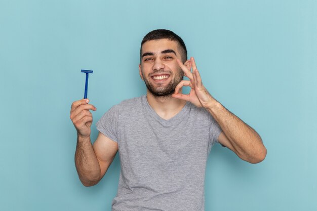 Vista frontal hombre joven en camiseta gris sosteniendo navaja y sonriendo en el escritorio azul barba de afeitar color de espuma de cabello masculino