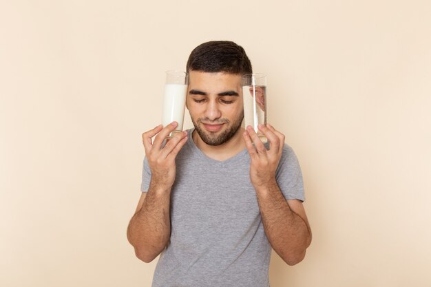 Vista frontal del hombre joven en camiseta gris con vasos de agua, leche en beige