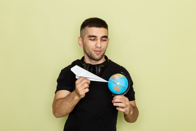 Vista frontal del hombre joven en camiseta negra con avión de papel y globo terráqueo en verde