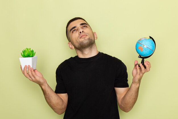 Vista frontal del hombre joven en camiseta negra sosteniendo una pequeña planta verde y un pequeño globo mientras piensa en verde