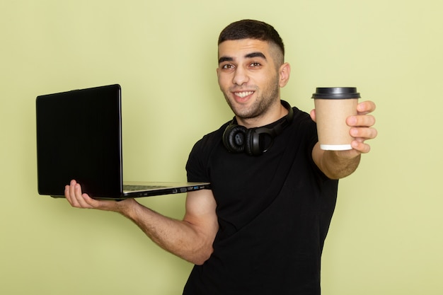 Vista frontal del hombre joven en camiseta negra usando laptop sosteniendo una taza de café con una sonrisa en verde