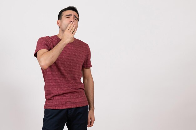 Vista frontal del hombre joven en camiseta de pie y bostezando sobre fondo blanco.