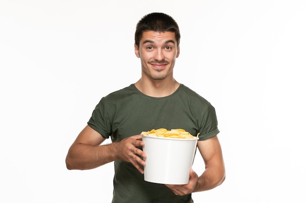 Vista frontal del hombre joven en camiseta verde sosteniendo la canasta con patatas cips y sonriendo en la pared blanca cine de película de disfrute solitario