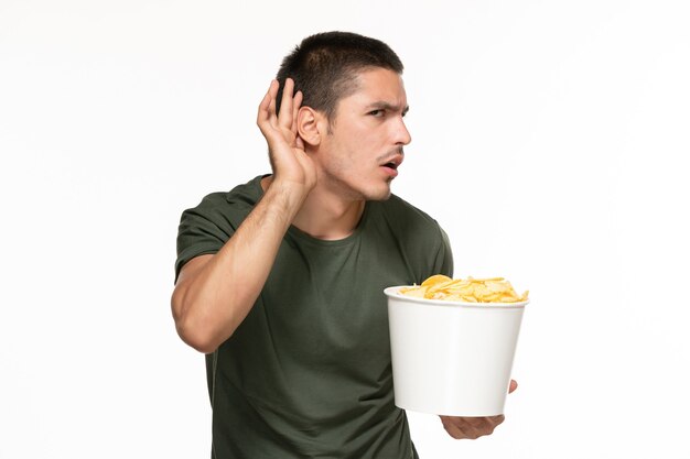 Vista frontal del hombre joven en camiseta verde sosteniendo una canasta con patatas cips y tratando de escuchar en la pared blanca película de cine de película de disfrute solitario