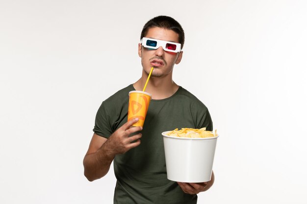 Vista frontal hombre joven en camiseta verde sosteniendo cips de patata y bebiendo refrescos en gafas de sol d en película de pared blanca película solitaria masculina