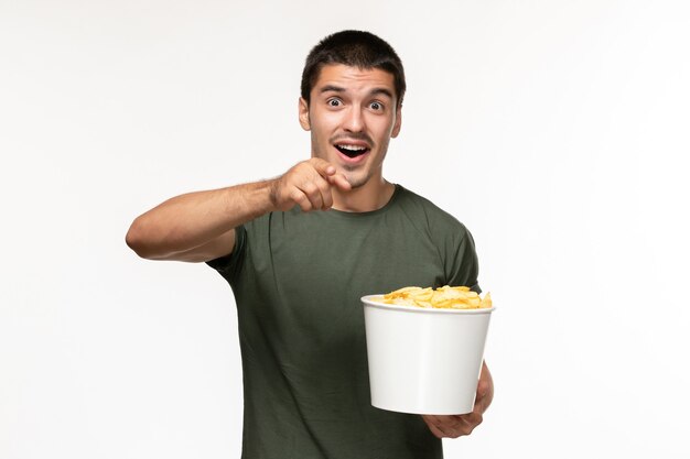 Vista frontal del hombre joven en camiseta verde sosteniendo cips de patata y viendo la película en la pared blanca clara película solitaria película cine