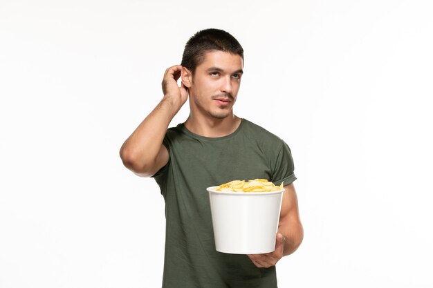 Vista frontal del hombre joven en camiseta verde sosteniendo cips de patata y viendo la película en la pared blanca película solitaria película cine