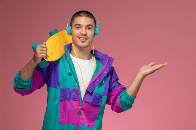 Vista frontal del hombre joven en capa colorida escuchando música con patineta en el escritorio rosa macho