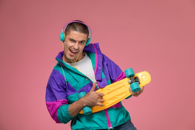 Vista frontal del hombre joven en capa colorida escuchando música con patineta en escritorio rosa