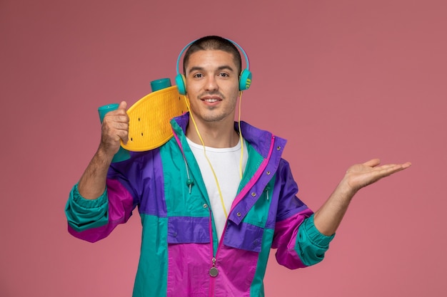 Vista frontal del hombre joven en capa colorida escuchando música con patineta en escritorio rosa