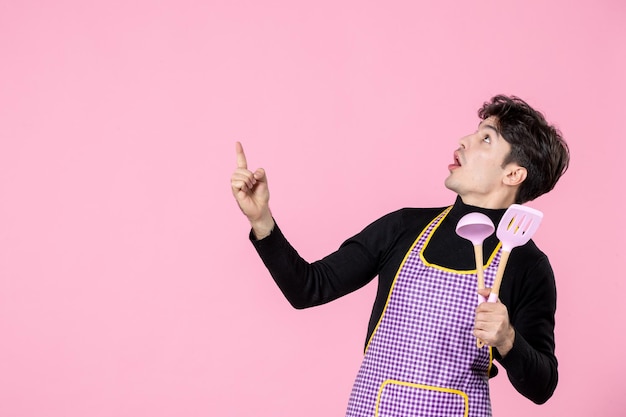 Vista frontal hombre joven en capa sosteniendo cucharas rosadas sobre fondo rosa masa profesión cocina uniforme horizontal jefe cocina trabajo color trabajador
