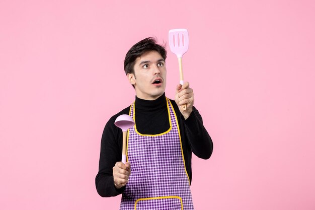 Vista frontal hombre joven en capa sosteniendo cucharas sobre fondo rosa color comida cocina uniforme trabajo de profesión de cocina horizontal