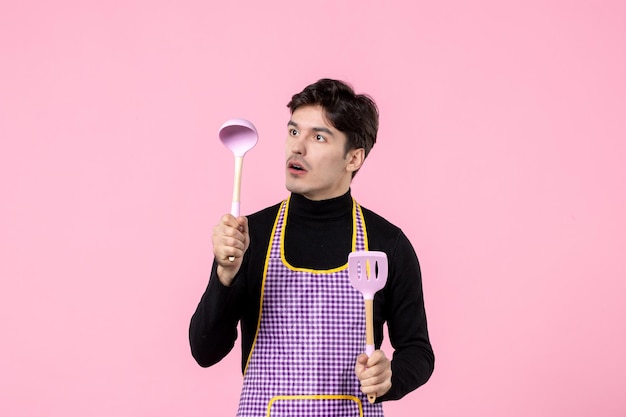 Vista frontal hombre joven en capa sosteniendo cucharas sobre fondo rosa color comida cocina uniforme trabajo de profesión de masa horizontal