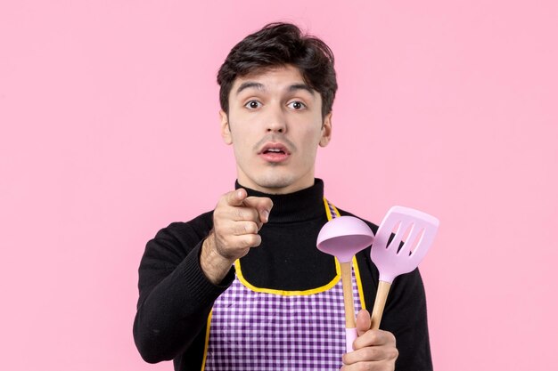 Vista frontal hombre joven en capa sosteniendo cucharas sobre fondo rosa profesión cocinar comida trabajo horizontal cocina color comida masa uniforme