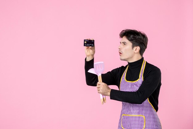 Vista frontal hombre joven en capa con tarjeta bancaria y cucharas sobre fondo rosa trabajo uniforme horizontal trabajador de cocina color cocina profesión jefe dinero