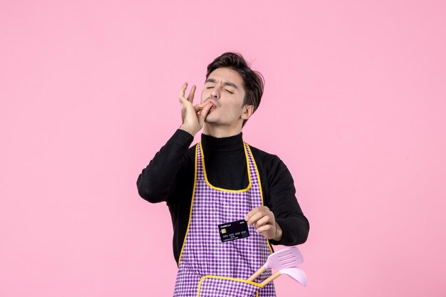 Vista frontal hombre joven en capa con tarjeta bancaria negra sobre fondo rosa profesión de trabajo jefe de cocina uniforme trabajador de cocina color horizontal