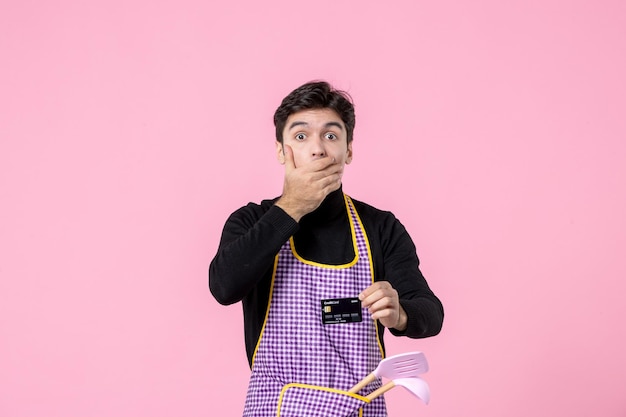 Vista frontal hombre joven en capa con tarjeta bancaria negra sobre fondo rosa trabajo profesión jefe de cocina trabajador uniforme color dinero cocina