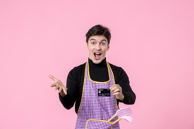 Vista frontal hombre joven en capa con tarjeta bancaria negra sobre fondo rosa trabajo profesión jefe de cocina trabajador uniforme dinero cocina horizontal