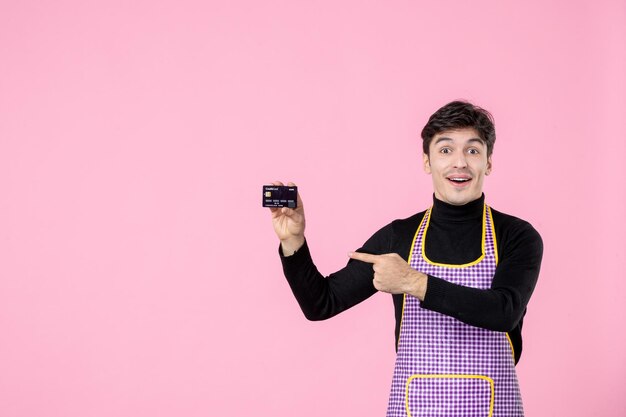 Vista frontal hombre joven en capa con tarjeta bancaria sobre fondo rosa color uniforme trabajo cocina trabajador cocina profesión jefe